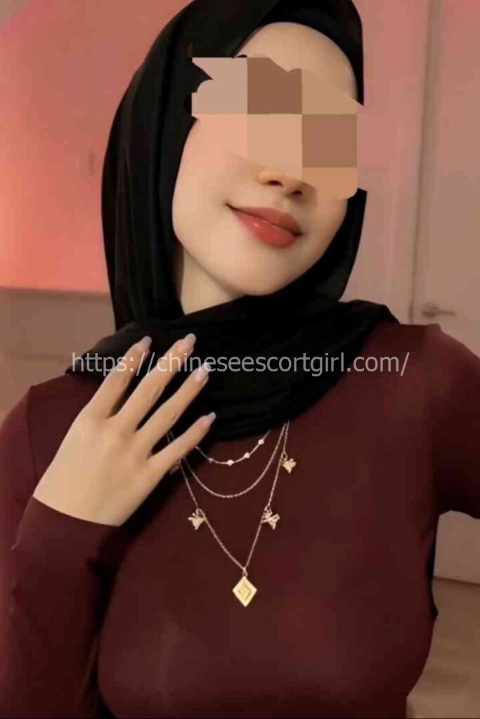 Young woman in hijab and maroon top, wearing layered necklaces.